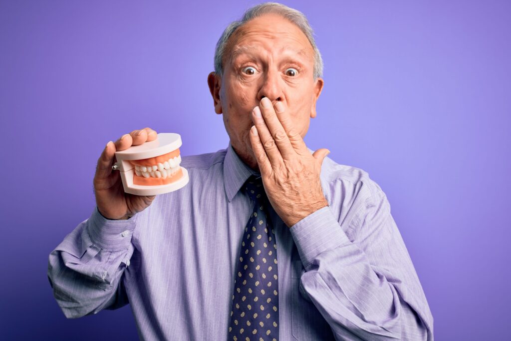 Man in purple shirt with hand over mouth holding dentures looking shocked