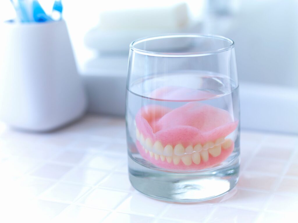 Full set of dentures in a glass of clear fluid on tiled counter